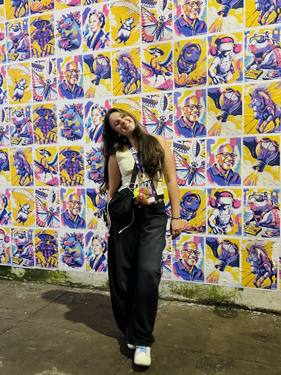 Camila Maia sorrindo em frente a um muro de pôsteres coloridos